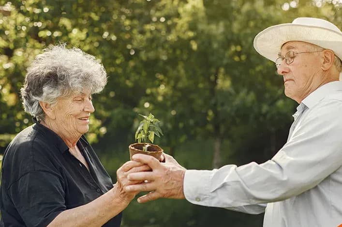 An older woman handing a potted plant to an older man in a garden