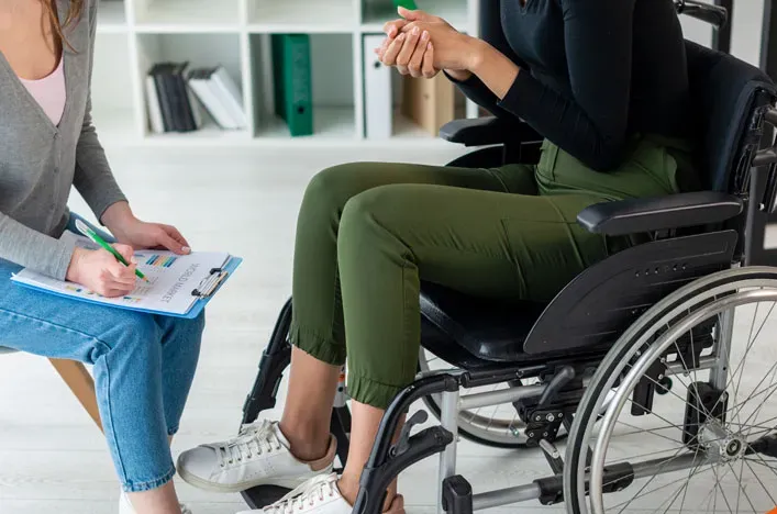 A care worker with a clipboard sitting beside a person in a wheelchair