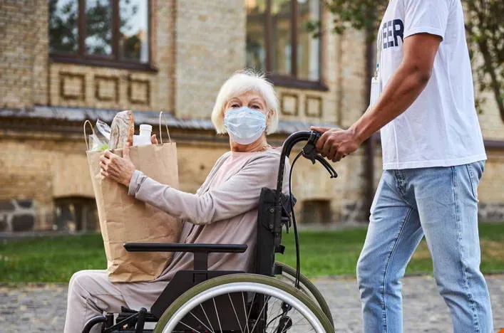 An older woman in a wheelchair holding a bag of groceries, with a helper beside her