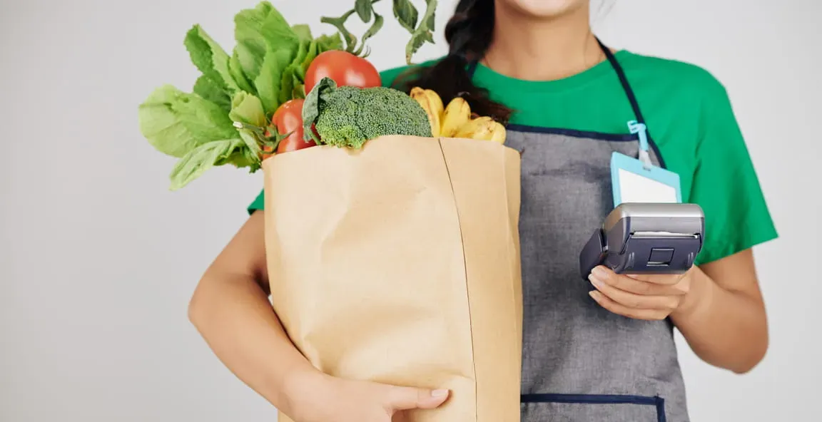 A grocery worker holding a full paper bag of vegetables and a card reader